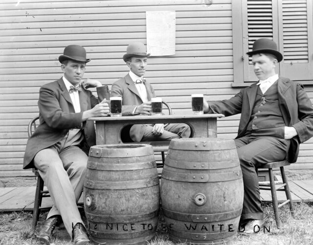 Black and white photograph of men in suits sitting around table holding beers with two barrels in front. Writing on photo says "Nice to have been waited on."