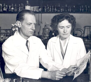 Carl and Gerty Cori in their lab, wearing white lab coats. Carl points to a document with a pencil. There are beakers and bottles in the background.