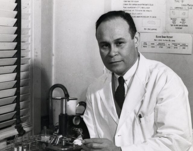 Charles Drew is seated at a counter wearing a lab coat and touching a microscope