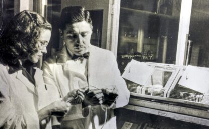 The Frees wearing lab coats and standing in a research lab. Alfred holds what seems to be an animal test subject while Helen injects something.