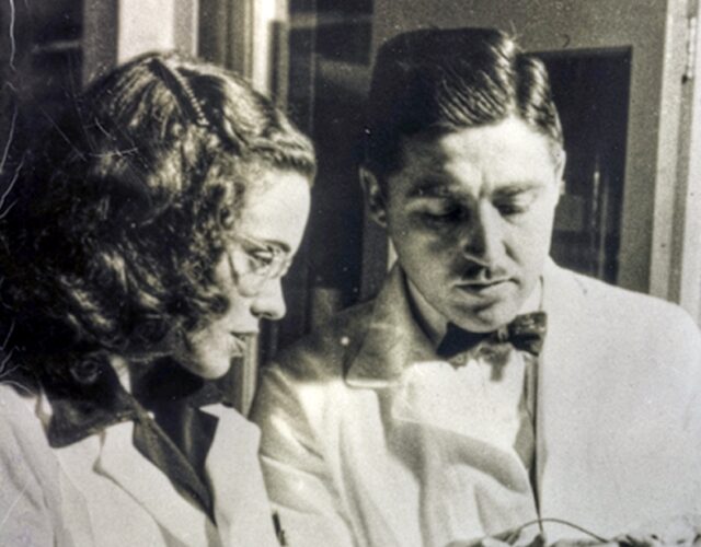 The Frees wearing lab coats and standing in a research lab. Alfred holds what seems to be an animal test subject while Helen injects something.