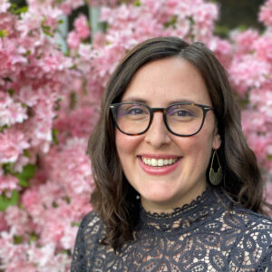 Caitlin in a dark grey lace shirt with hair down and glasses on in front of a pink azalea bush
