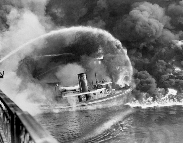 Black and white photograph of a boat on fire in a river and a small hose attempting to extinguish it.