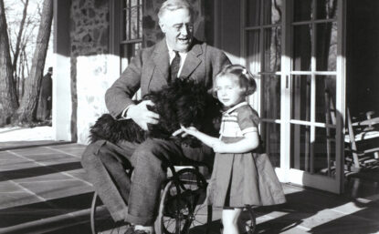 Black and white photograph of US president Franklin Roosevelt in wheelchair, holding a dog in his lap, with a young girl next to him.