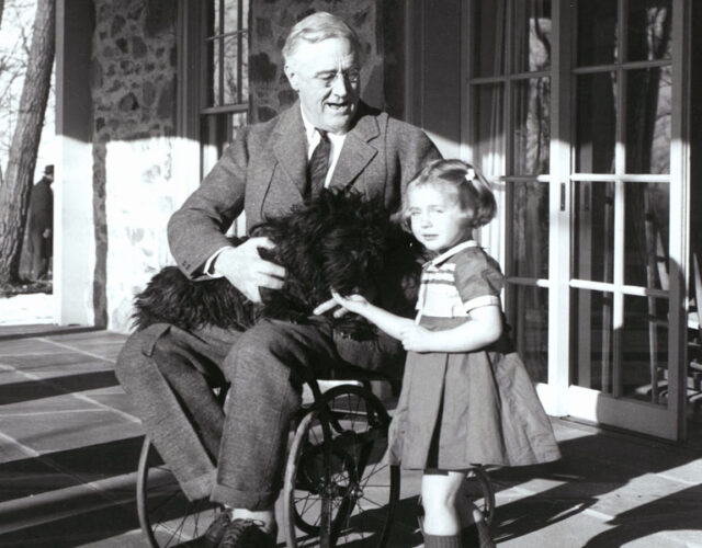 Black and white photograph of US president Franklin Roosevelt in wheelchair, holding a dog in his lap, with a young girl next to him.