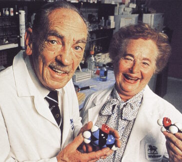Hitchings and Elion in a lab wearing lab coats and smiling. They both hold black, blue, red, and white plastic models