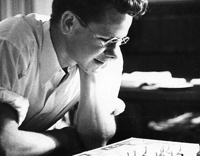 Black and white photograph of a young Gordon Gould looking down at a machine. He is wearing glasses and a white button down shirt.