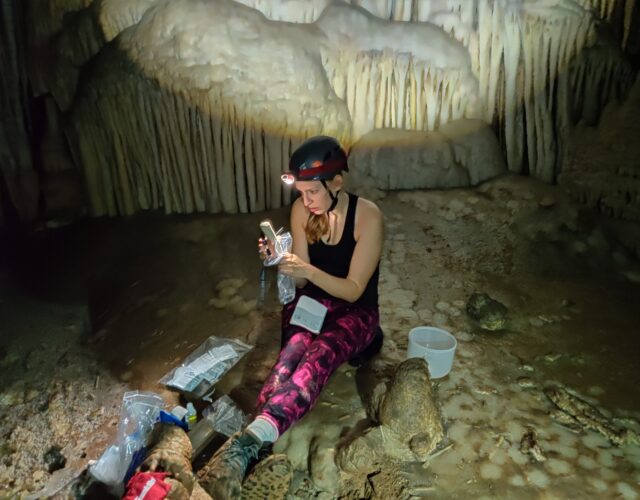 Photograph of scientist Gabriela Serrato Marks wearing a headlamp in a cave, surrounded by samples in plastic bags.