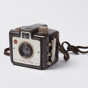 Kodak brand Brownie Holiday Flash camera; Dakon plastic lens with rotary shutter; molded brown and tan Bakelite body; three metal sockets on right side of the camera body meant for mounting flash bulb; braided cord carrying strap.