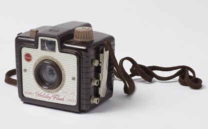 Kodak brand Brownie Holiday Flash camera; Dakon plastic lens with rotary shutter; molded brown and tan Bakelite body; three metal sockets on right side of the camera body meant for mounting flash bulb; braided cord carrying strap.