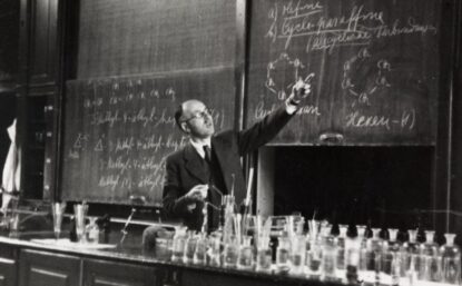 A black and white photo of a man wearing glasses in a suite and tie pointing at a chalk board. There is a counter filled with glass vessels in the foreground.