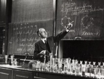 A black and white photo of a man wearing glasses in a suite and tie pointing at a chalk board. There is a counter filled with glass vessels in the foreground.