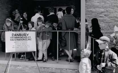 Black and white photograph of adults and children waiting to get inside a building. A sign reads "Danbury German measles vaccination clinic".