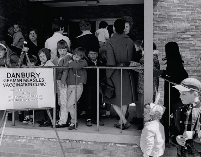 Black and white photograph of adults and children waiting to get inside a building. A sign reads "Danbury German measles vaccination clinic".