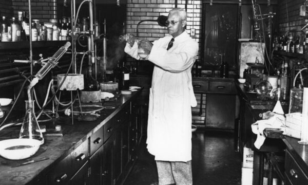 St. Elmo Brady in a chemistry lab at Fisk University. He is pouring a liquid from one beaker into another. There are counters of lab equipment surrounding him.