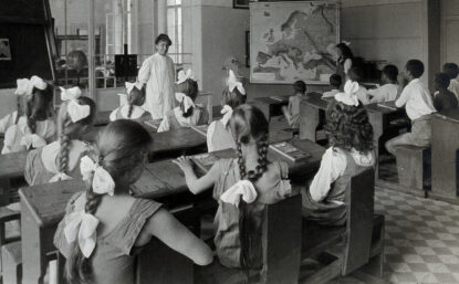 1920s black and white photograph of children learning geography in a classroom. Girls and boys are seated on different sides of the room.