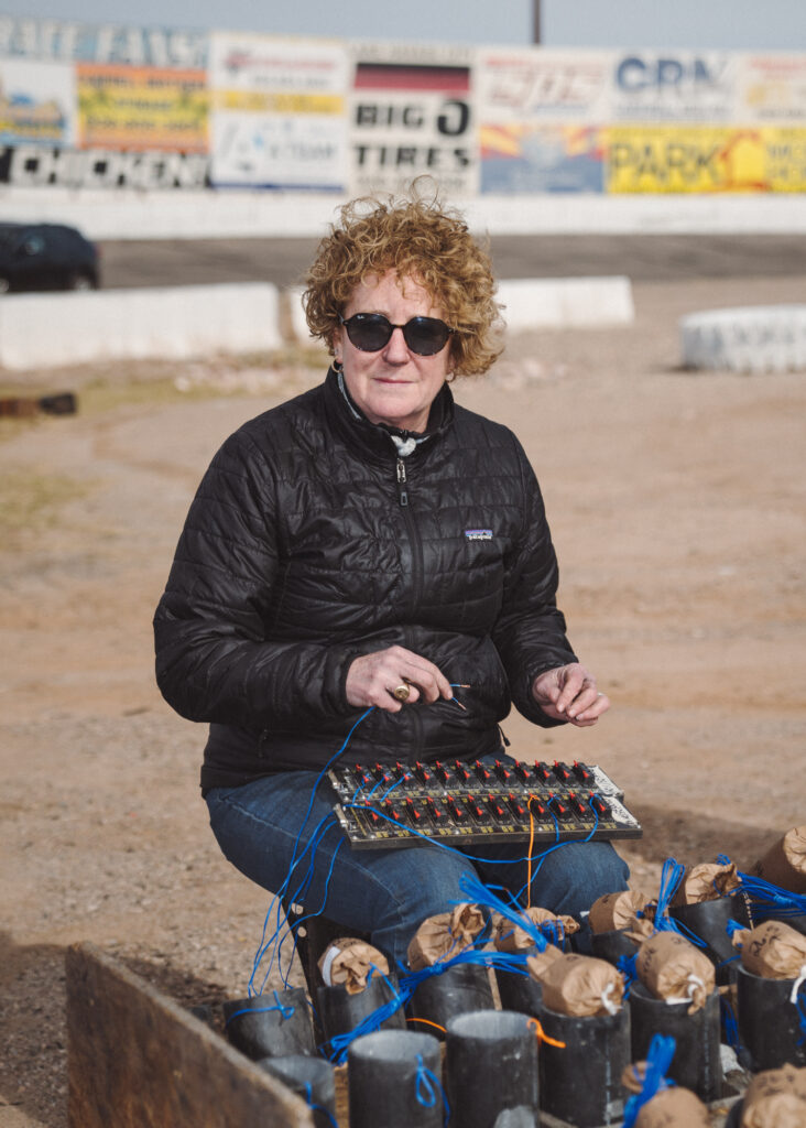 Woman seated outdoors holding electrical panel connected to pyrotechnical charges