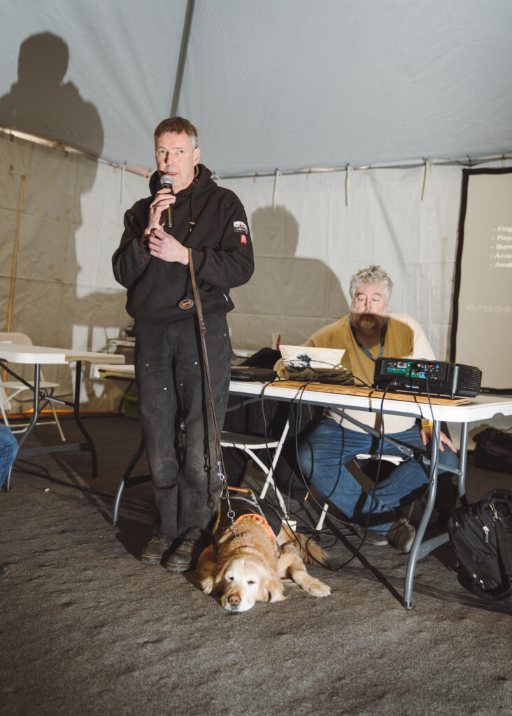 Man standing and holding a microphone inside a tent with a dog at his feet