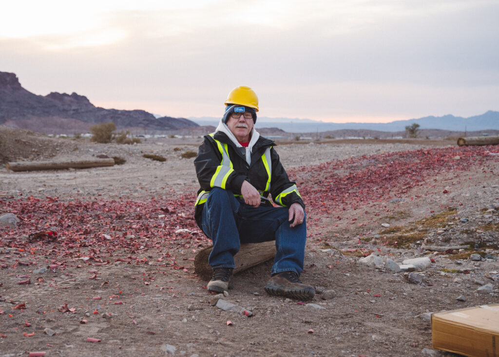 Older man in hard hat seated on log in desert landscape