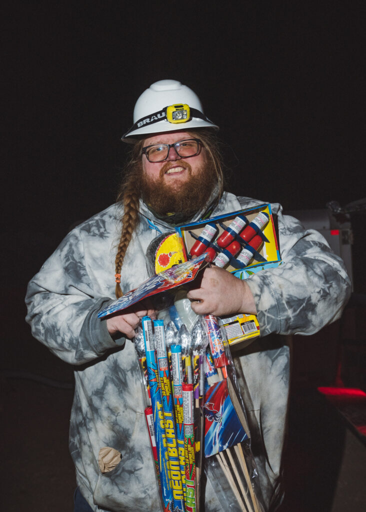 Bearded man standing in dark, wearing hard hat holding a lot of fireworks 