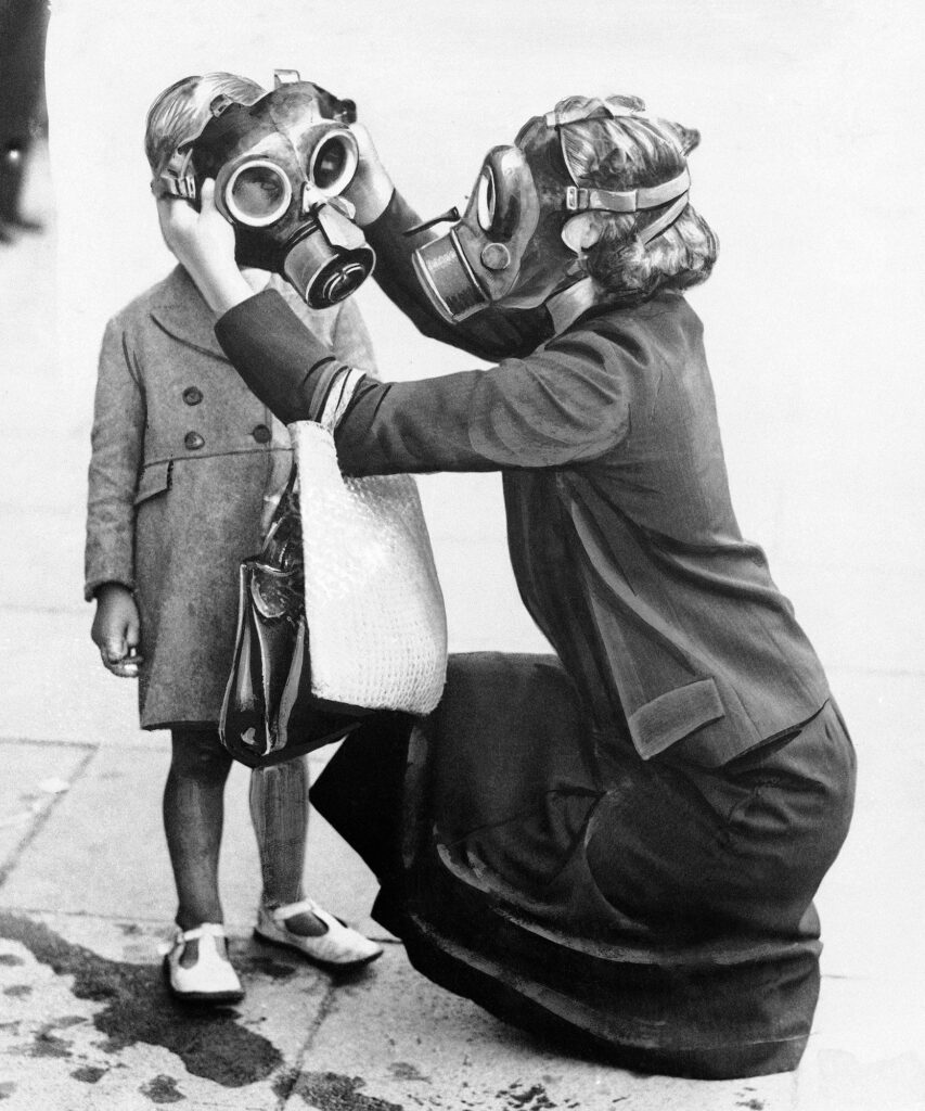 Woman wearing gas mask fits one on a child on a sidewalk