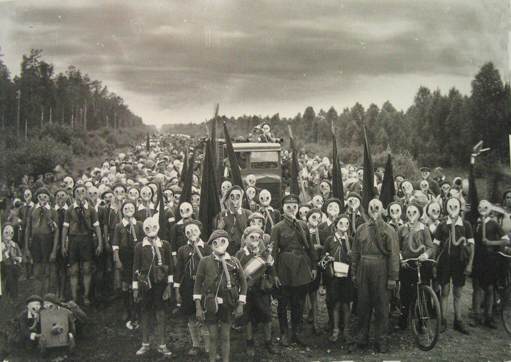Large group of children in clearing with military equipment and wearing gas masks