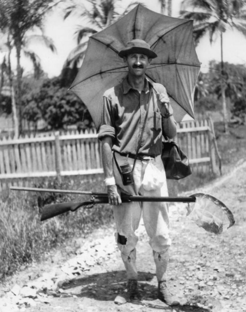 Black and white photo of a man holding a rifle and parasol in a tropical location