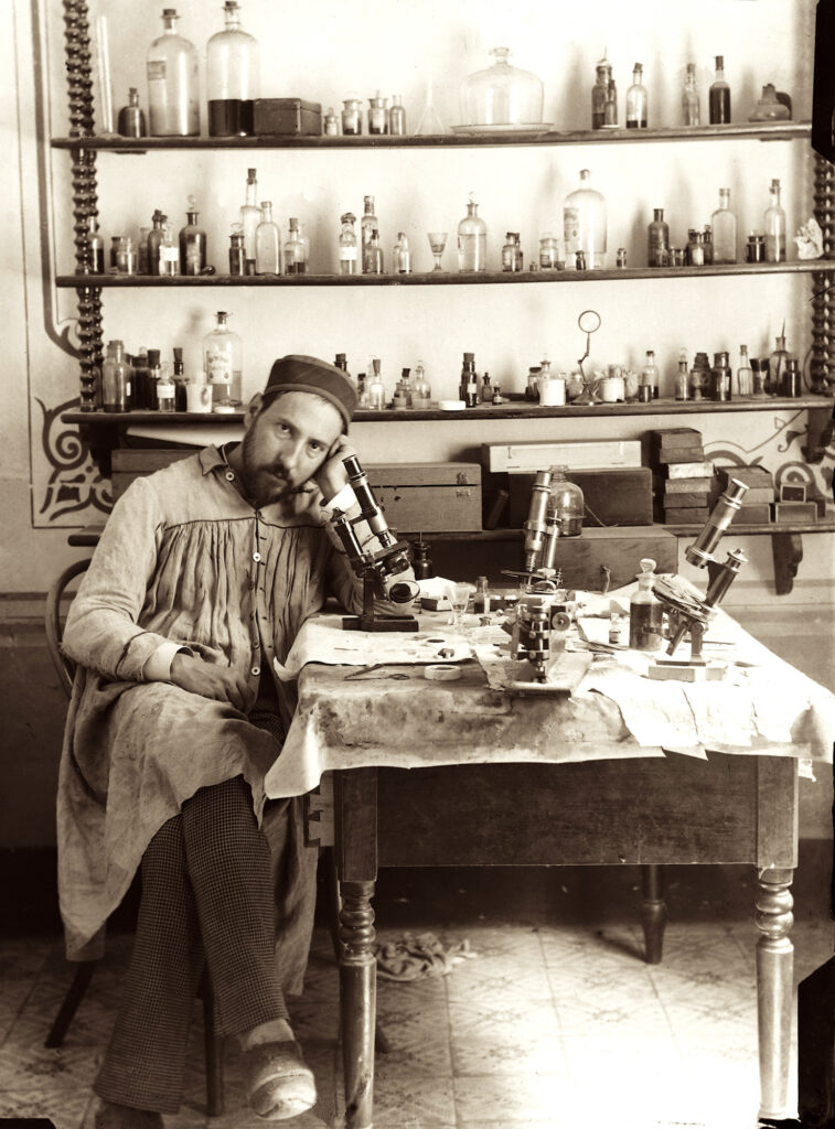 Photograph of man in lab apparel seated in front of microscope and samples 