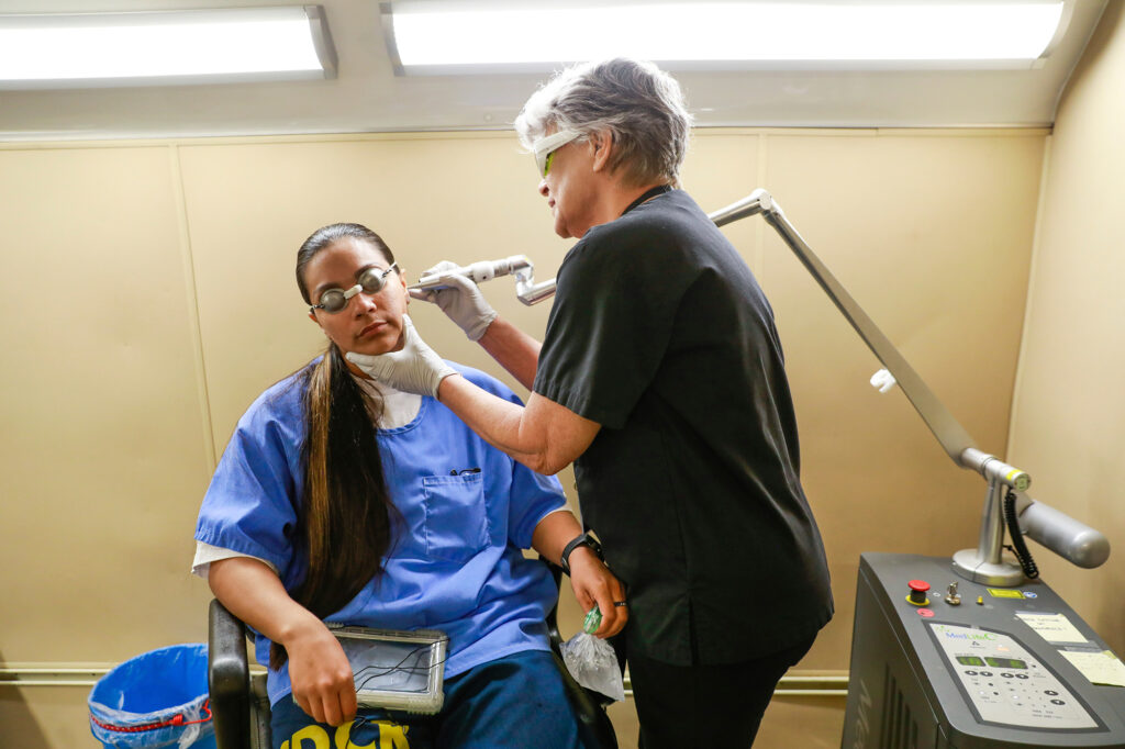 An older woman holding an articulated instrument to the face of a younger woman wearing eye protection