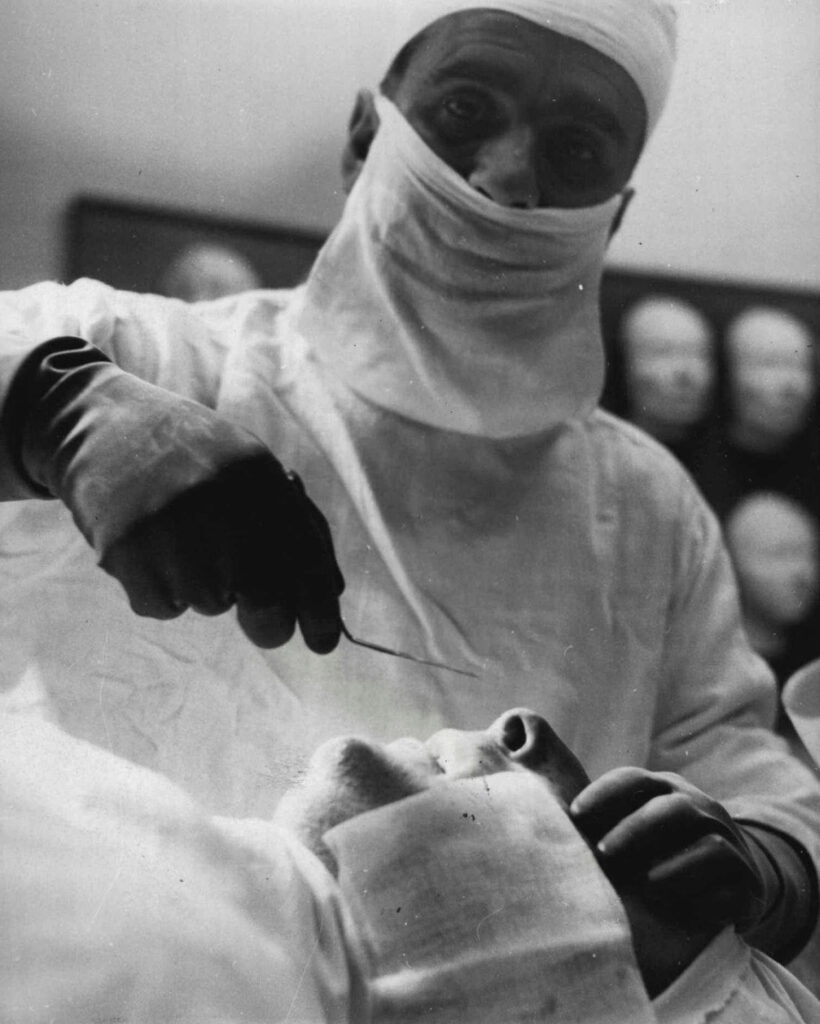 Black and white photo of a masked surgeon holding an instrument above the face of sedated patient