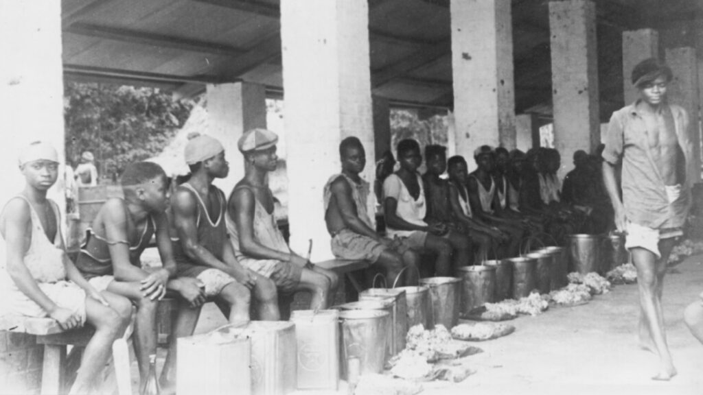 Large group of men and boys sitting on a covered bench with pails and hardened rubber in front of them