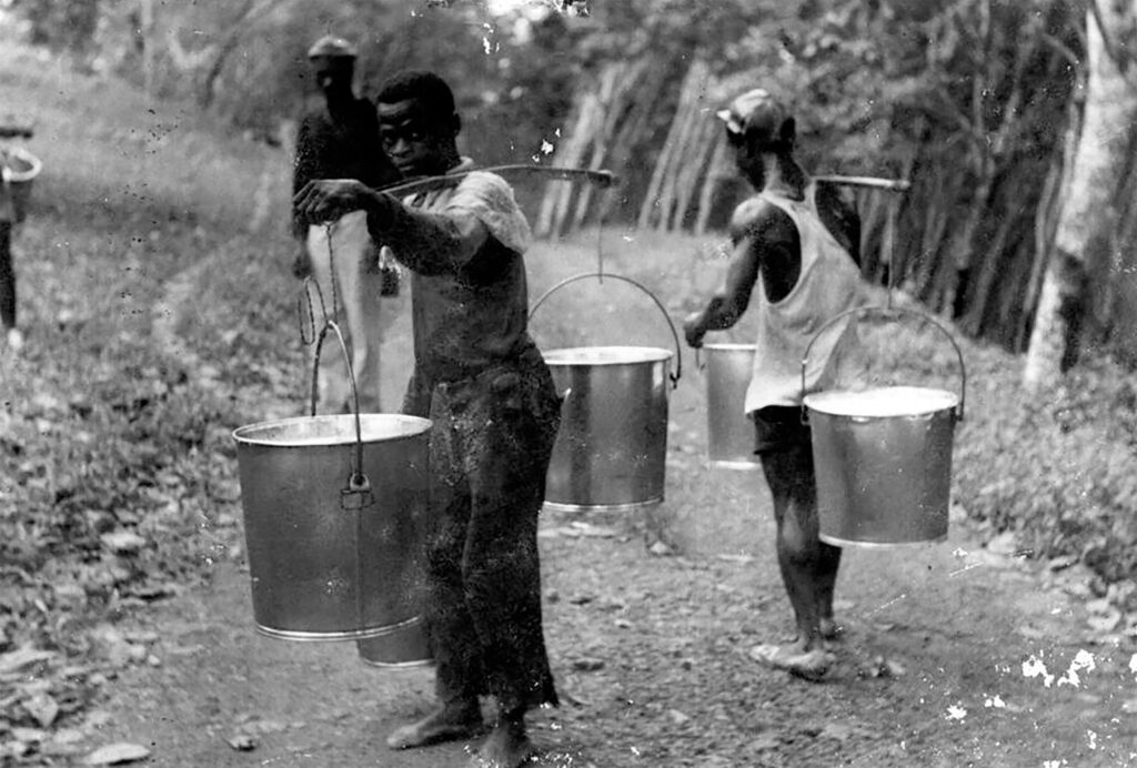 Men carrying large metal pails hanging from wood rods balanced over their shoulders