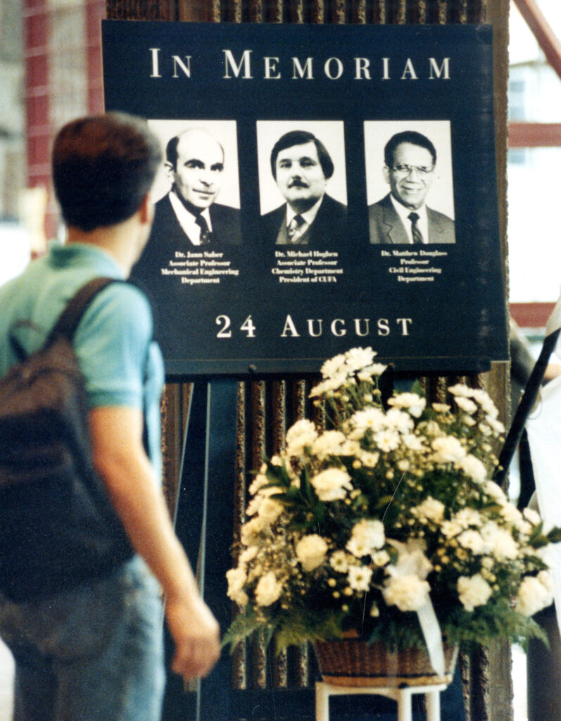 Color photo of man standing in front of a memorial board with three photo portraits and flowers 