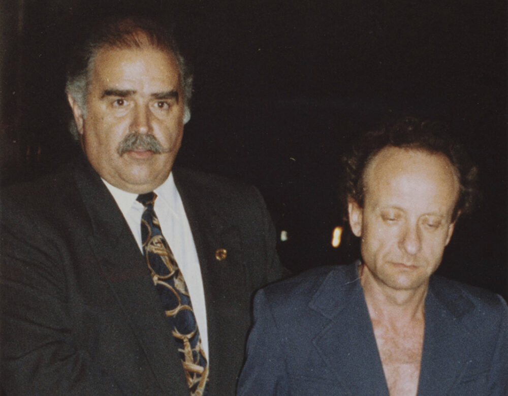 Color photo of two men in suits, one without a shirt, photographed walking in the dark