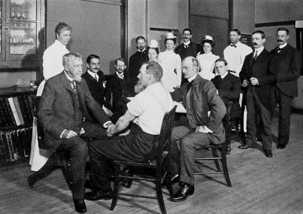 Photo of two men seated in a room with a small crowd of people looking on