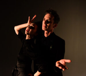 Ellia Bisker and Jeff Morris posing, leaning back with hands out, in a dark studio