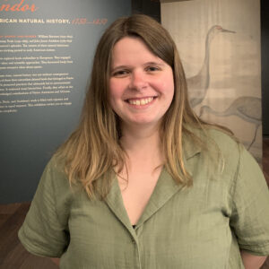 Ali Rospond in sage green, smiling, in front of exhibition display