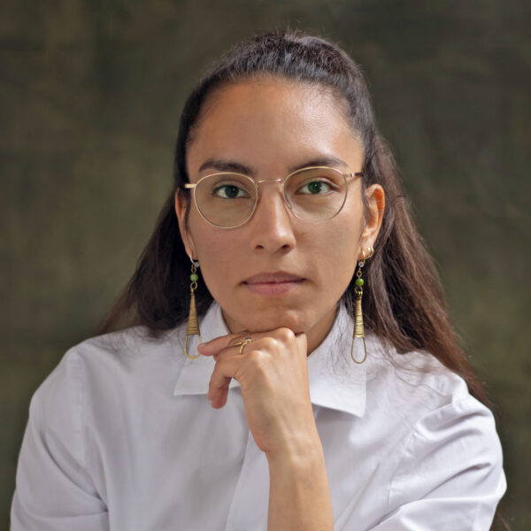 Studio portrait of Candida F. Sánchez Burmester wearing white dress shirt, glasses, long earrings, resting chin on hand