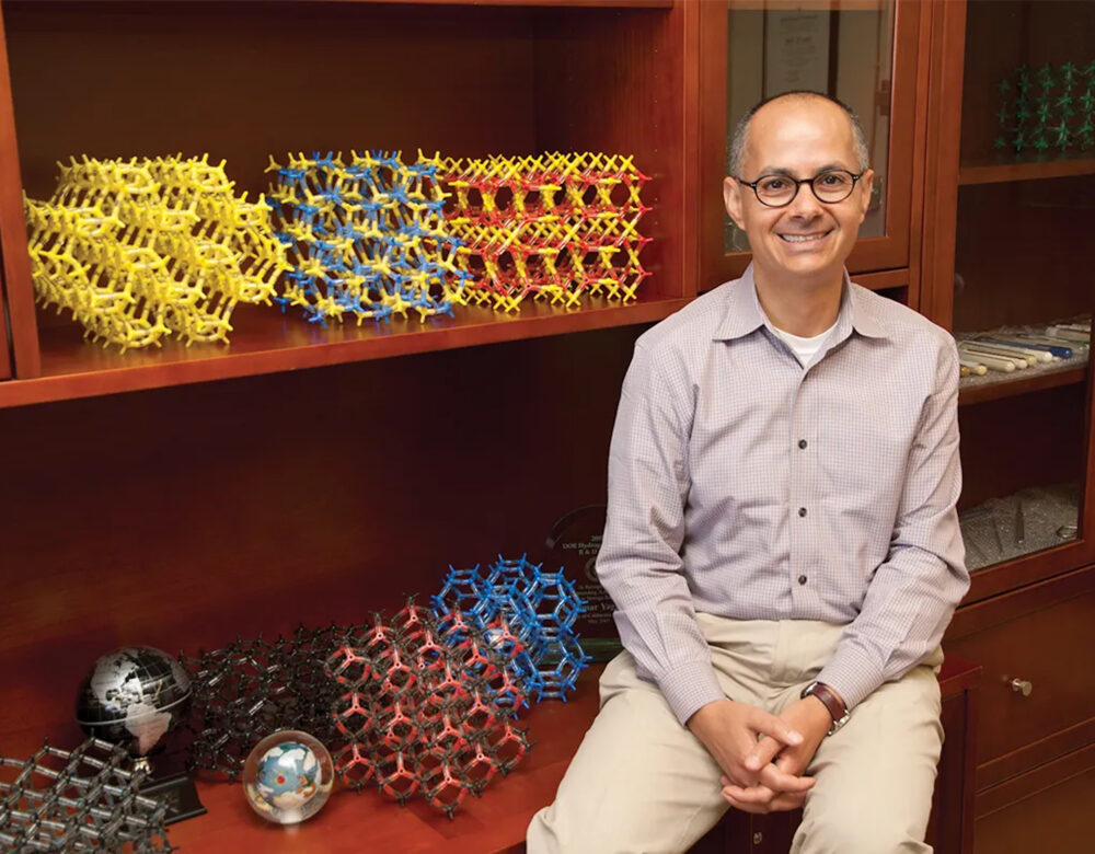 Omar Yaghi, smiling, hands clasped, seated by shelf with colorful molecular models and globes