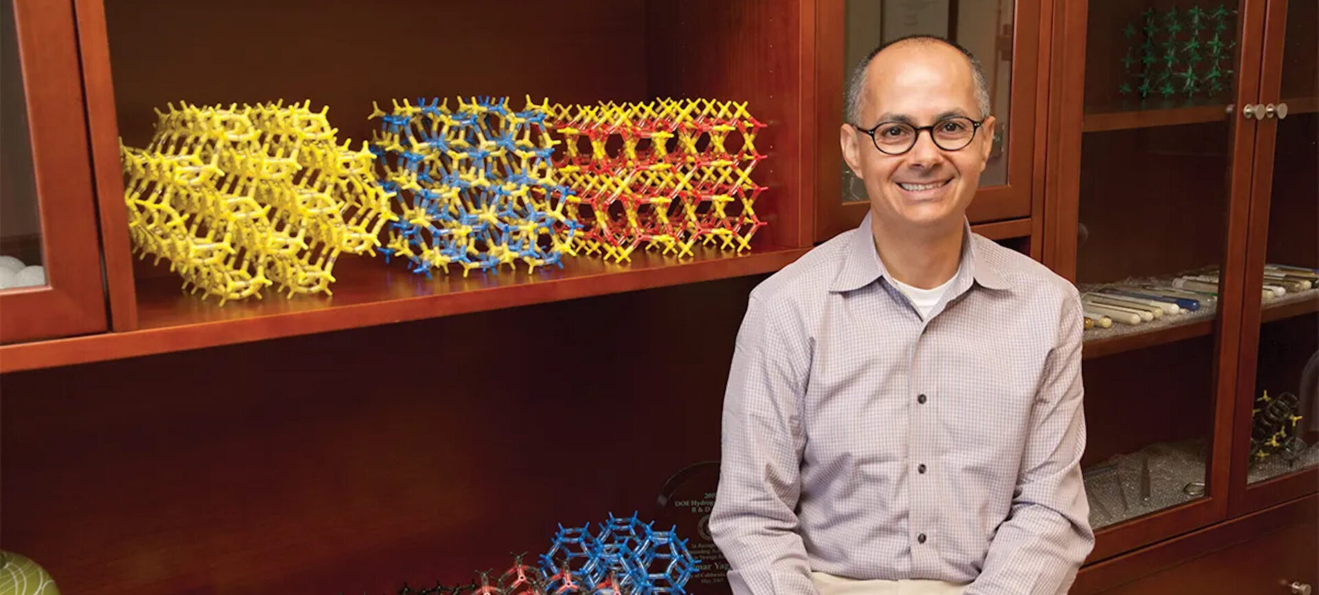 Omar Yaghi, smiling, hands clasped, seated by shelf with colorful molecular models and globes