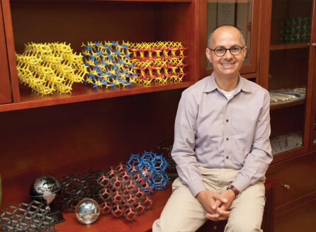 Omar Yaghi, smiling, hands clasped, seated by shelf with colorful molecular models and globes