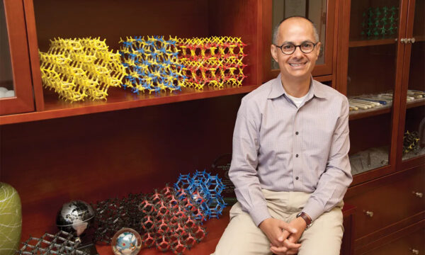 Omar Yaghi, smiling, hands clasped, seated by shelf with colorful molecular models and globes