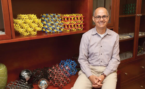 Omar Yaghi, smiling, hands clasped, seated by shelf with colorful molecular models and globes