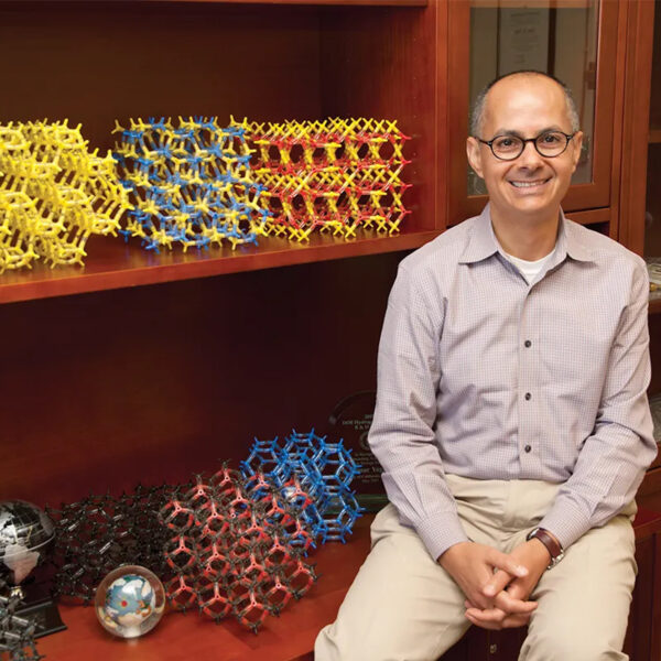 Omar Yaghi, smiling, hands clasped, seated by shelf with colorful molecular models and globes