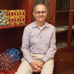 Omar Yaghi, smiling, hands clasped, seated by shelf with colorful molecular models