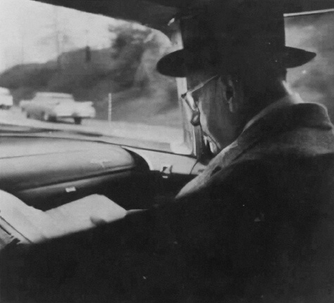 Black and white photo of an older man reading in a moving car, taken from the backseat