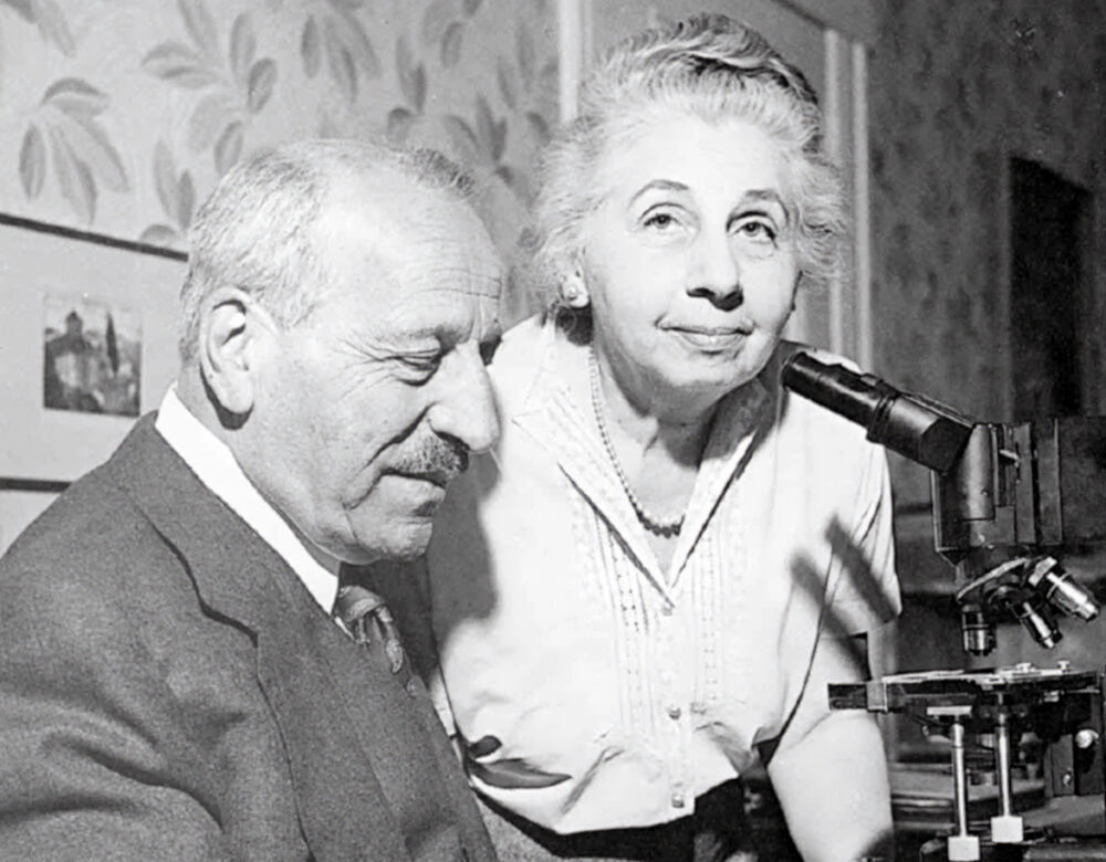 Black and white photo of a seated older man and standing older woman in front of a microscope