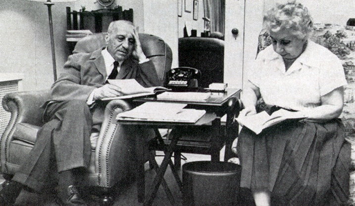 Black and white photo of a seated older man and older woman reading in a living room.