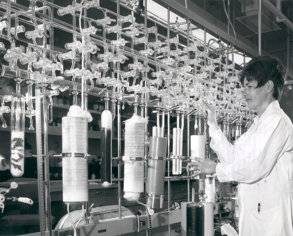 Black and white photo of a woman in lab coat adjusting a large set of chemical glass