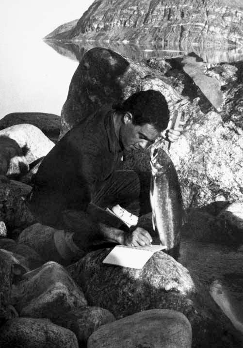 Black and white photo of man weighing fish by rocky shore